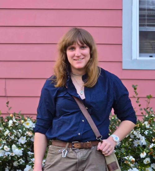 A woman in a button-down shirt and sidebag in front of a flower bush.